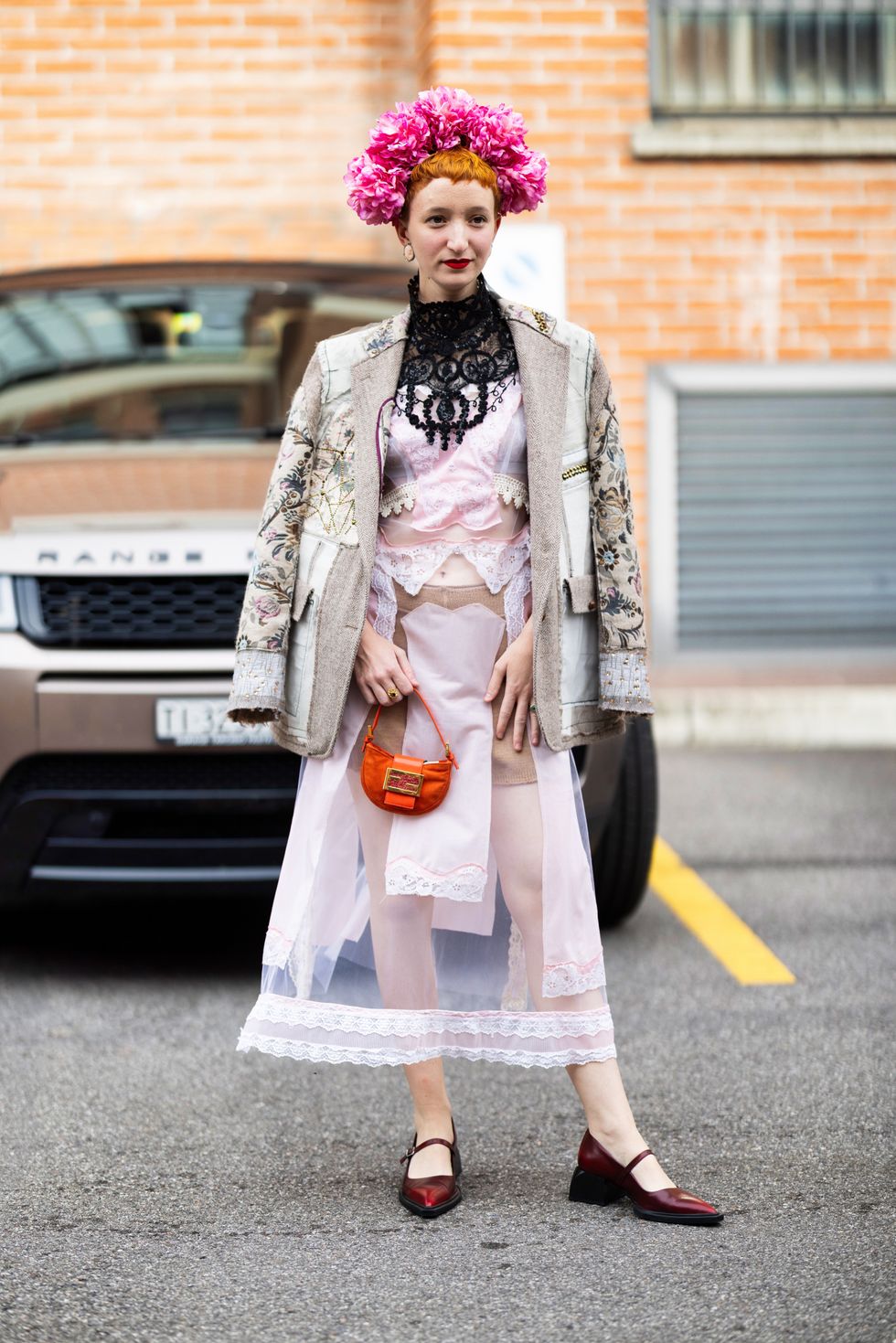 Judith Bradl is seen wearing an orange mini Fendi bag, red leather shoes, a pink flowers headpiece, a black lace collar, a multicolor beige printed blazer embellished with fabrics details and a see-through pink tulle dress embellished with lace details outside Antonio Marras show during the Milan Fashion Week
