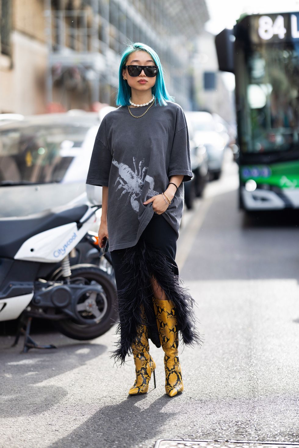 Margaret Zhang is seen wearing blue hair, black sunglasses, a pearl necklace, a gold necklace, yellow and black leather heels, an oversized grey t-shirt and a long black skirt embellished with feathers outside the Max Mara show during the Milan Fashion Week